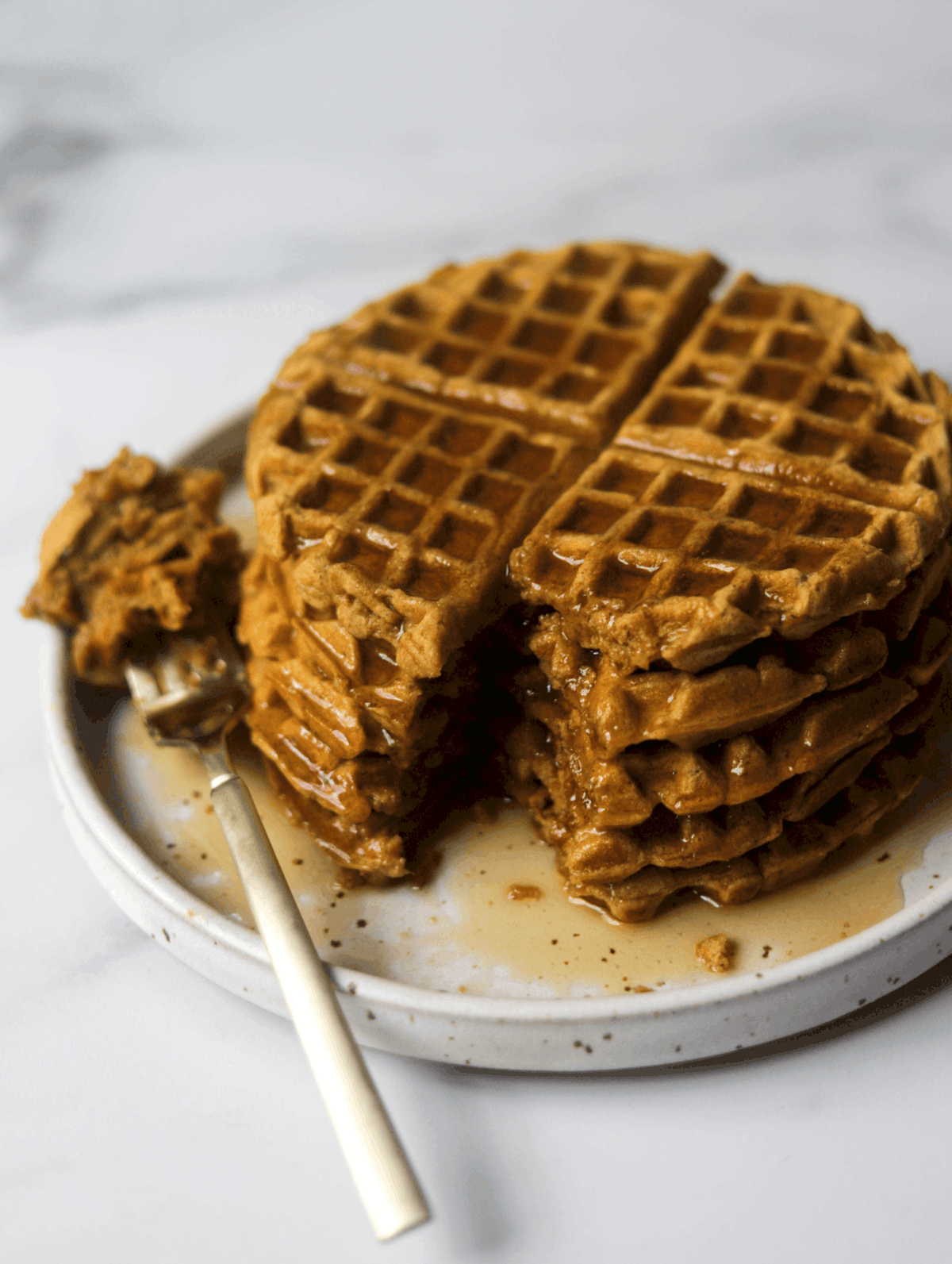 An angled shot of a plate of pumpkin waffles with maple syrup drizzled on top and a fork with a bite on it.