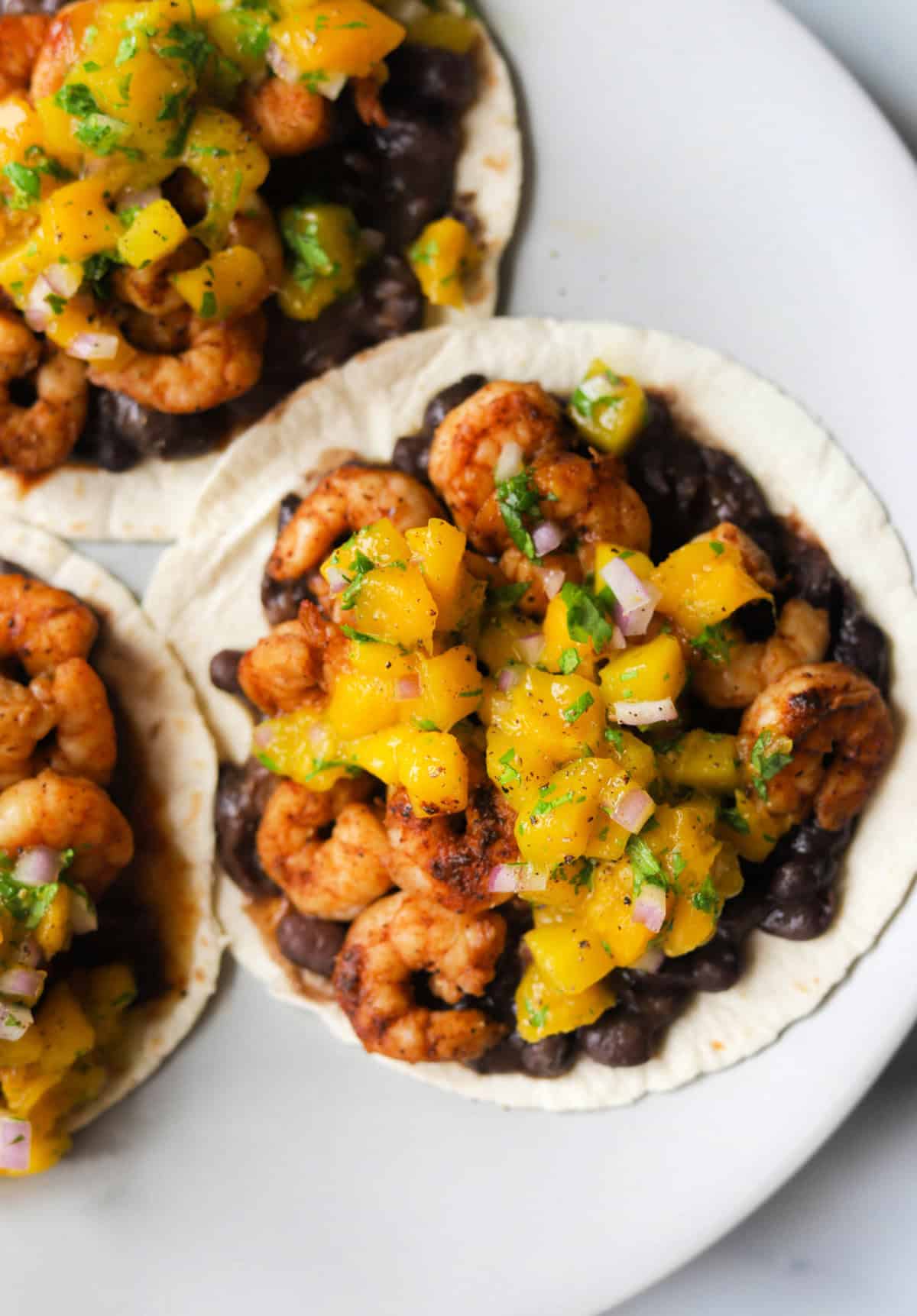 An overhead close up shot of spicy shrimp tacos with mango salsa on a light grey plate.