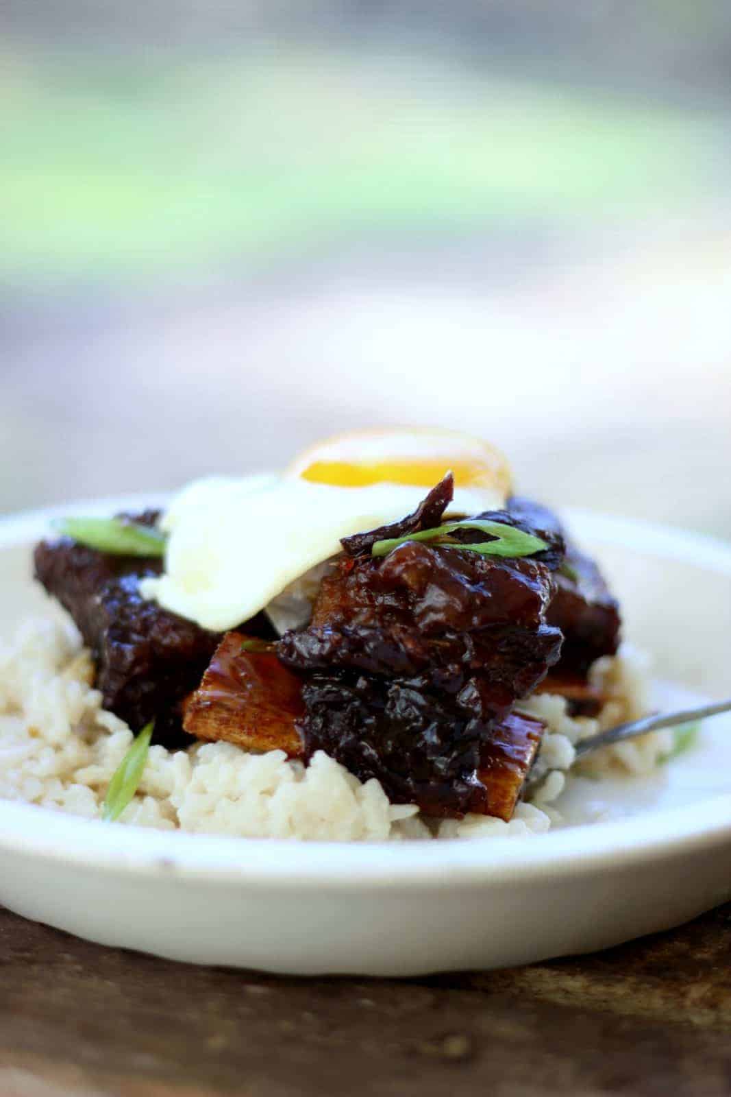 Side close-up shot of ginger beef short ribs on top of rice in a white bowl with an egg on top. 