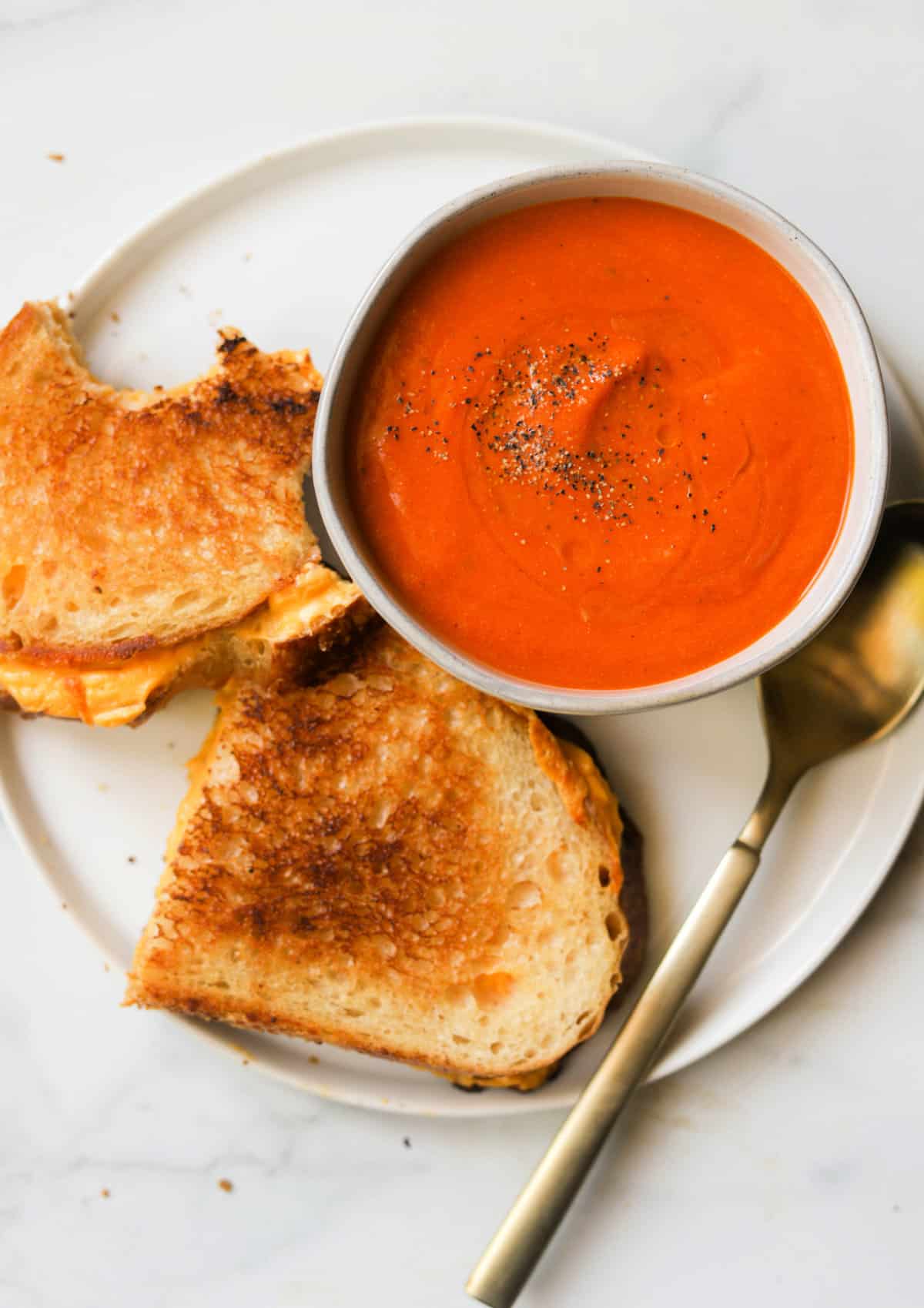 An overhead shot of a bowl of creamy tomato soup on top of a plate with a half eaten grilled cheese sandwich.