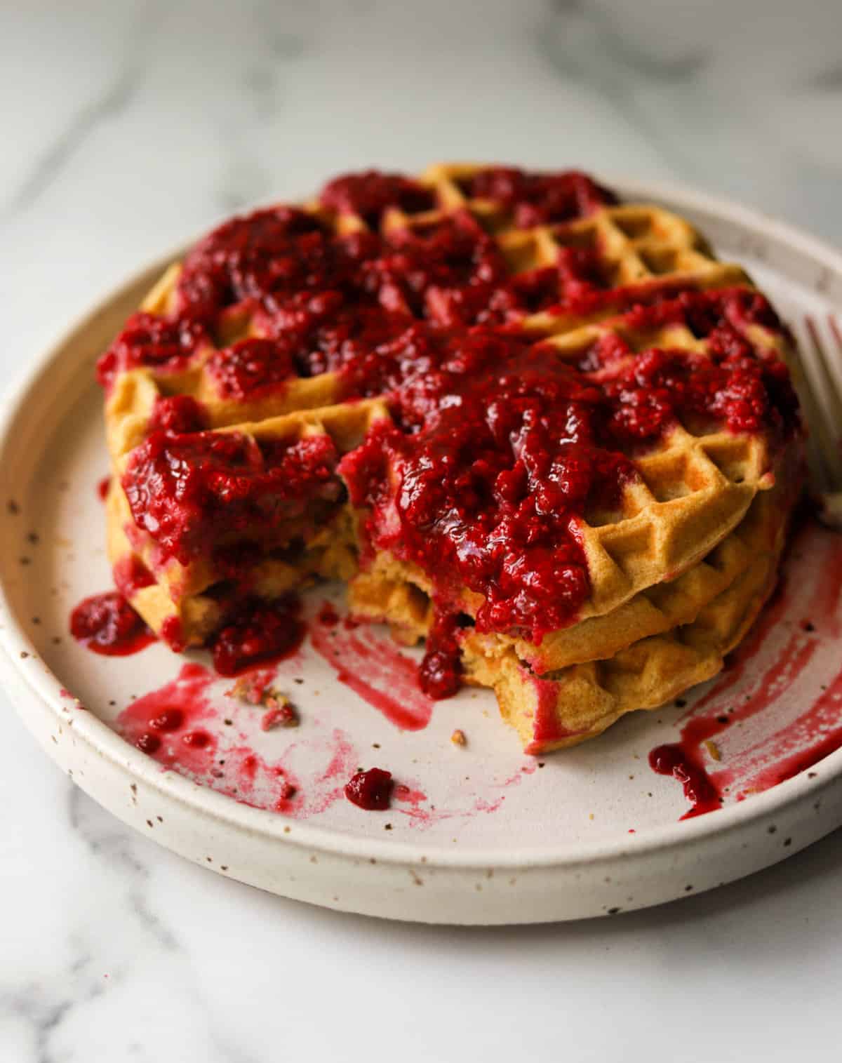 A side shot of a plate of lemon waffles with berry syrup with bites taken out of them.