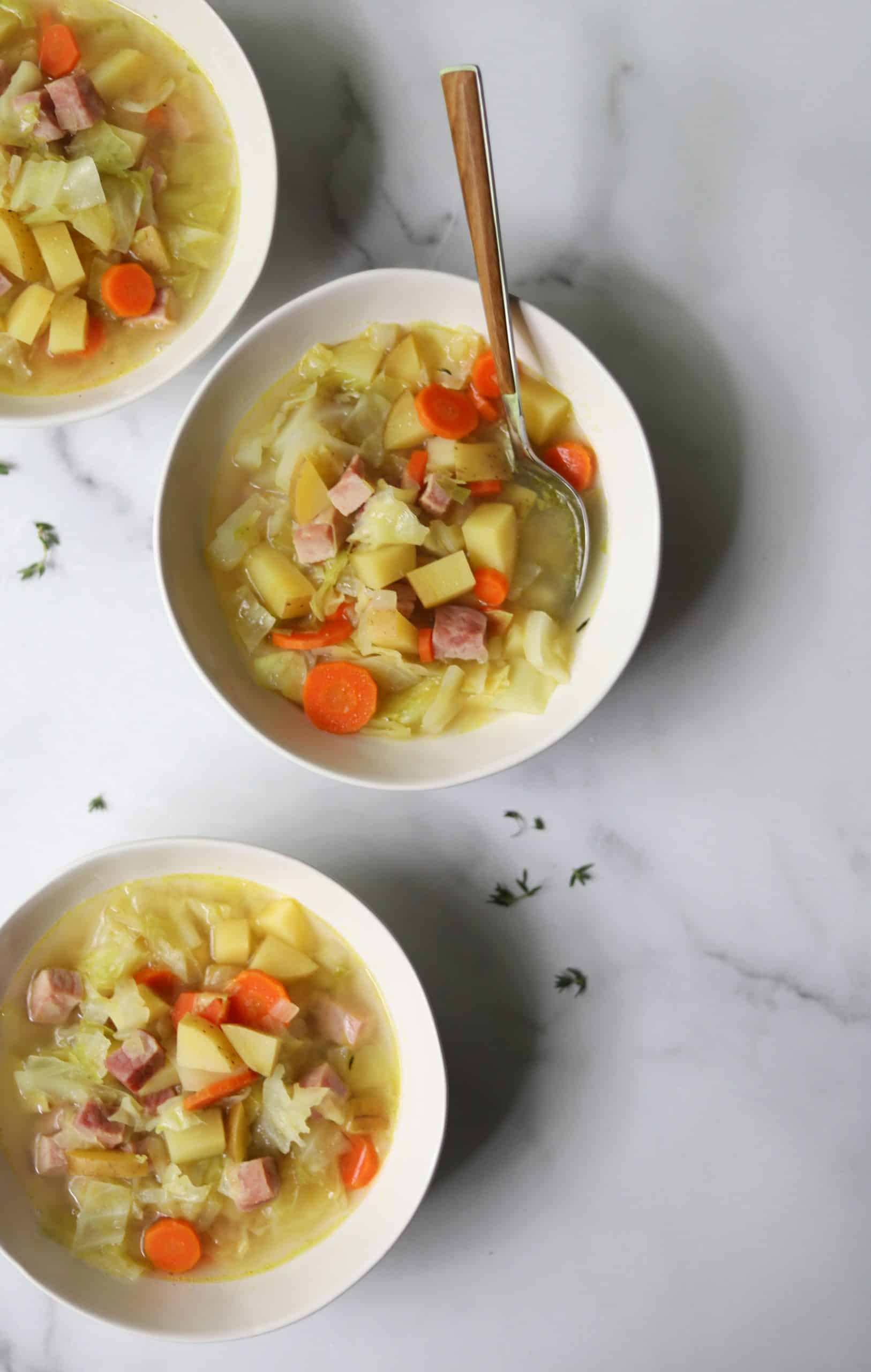 Three white bowls of ham, cabbage and potato soup on a white marble backdrop.