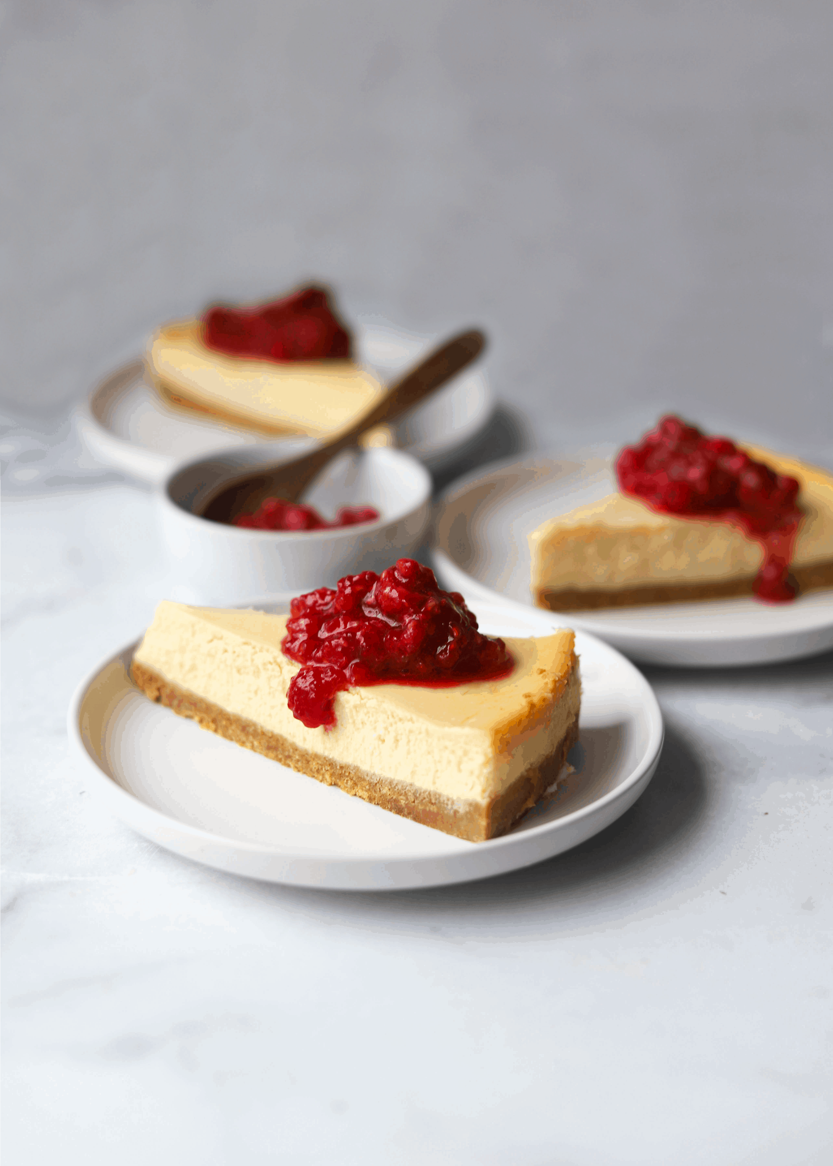 Three slices of cheesecake on white plates with a small bowl of raspberries.