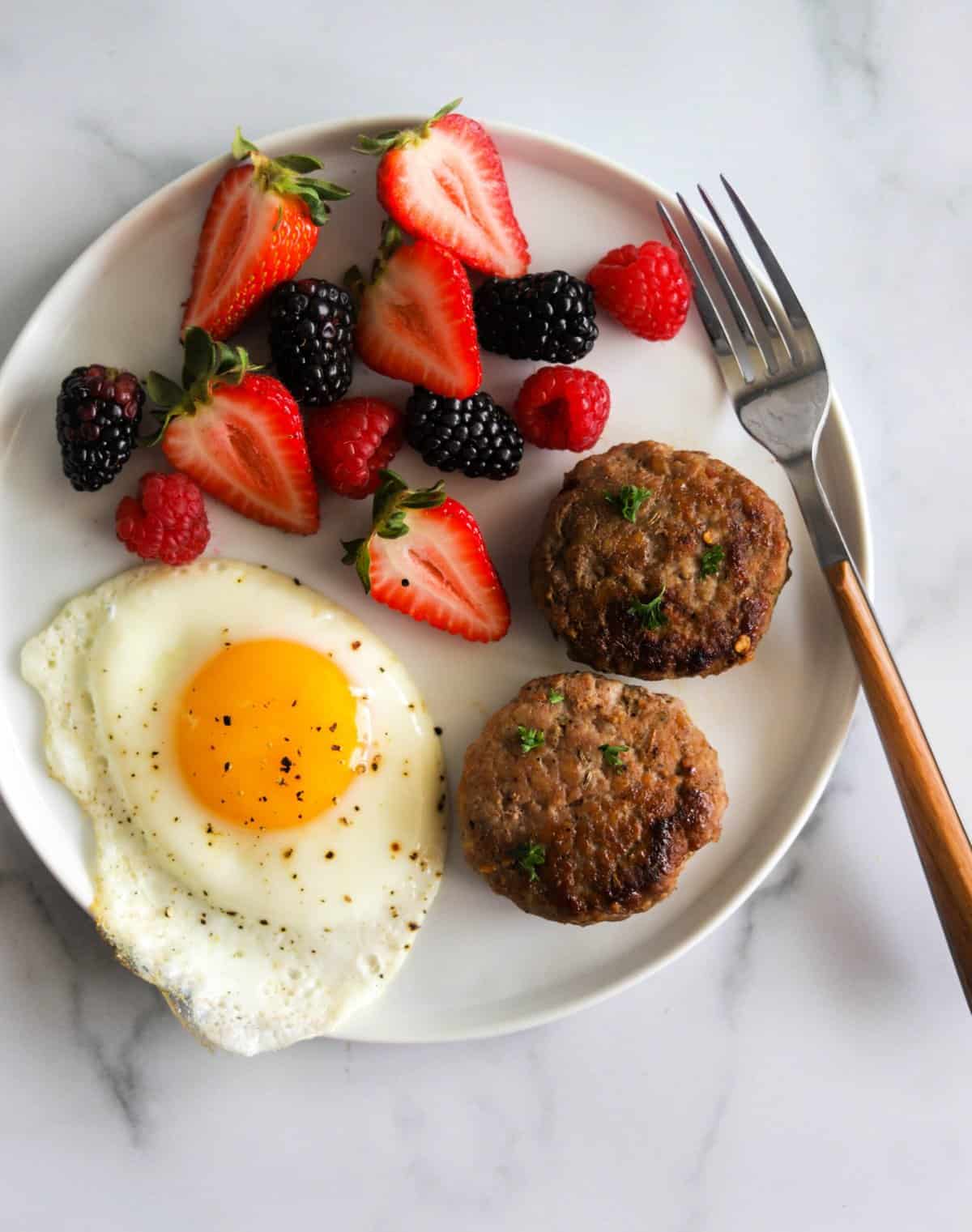 An overhead shot of a white plate with breakfast sausage, a fried egg and berries.