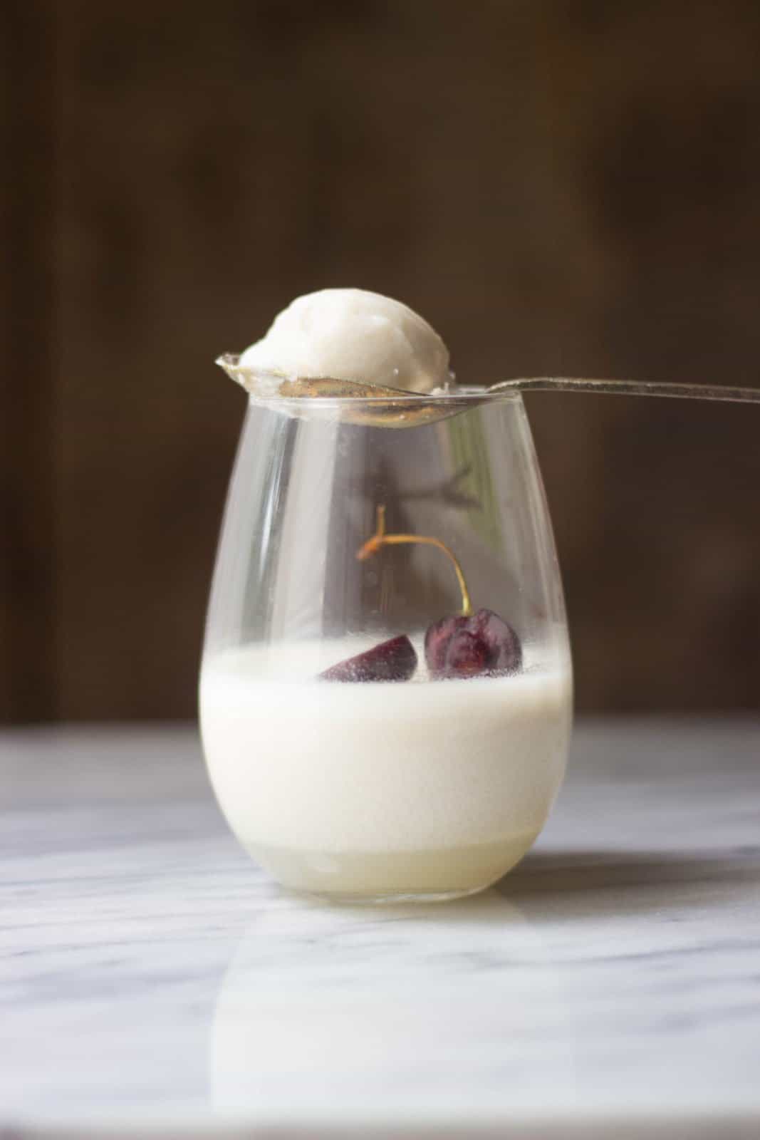 A bite of coconut milk panna cotta on a metal spoon balancing atop of a glass filled with panna cotta.