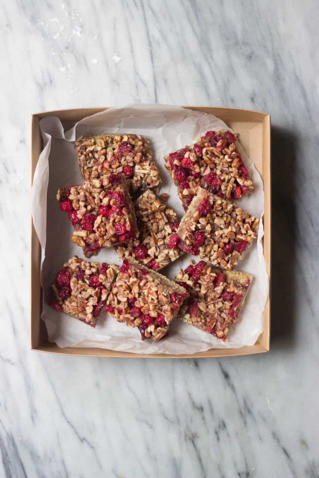 Overhead shot of eight cranberry pecan pie bars in a cardboard container with white parchment paper. 