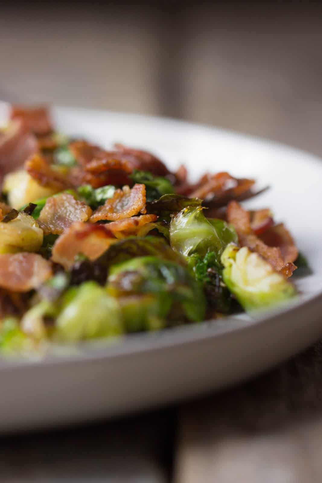 Side shot focused in on the cooked bacon on top of finished Brussels sprouts in a white plate.