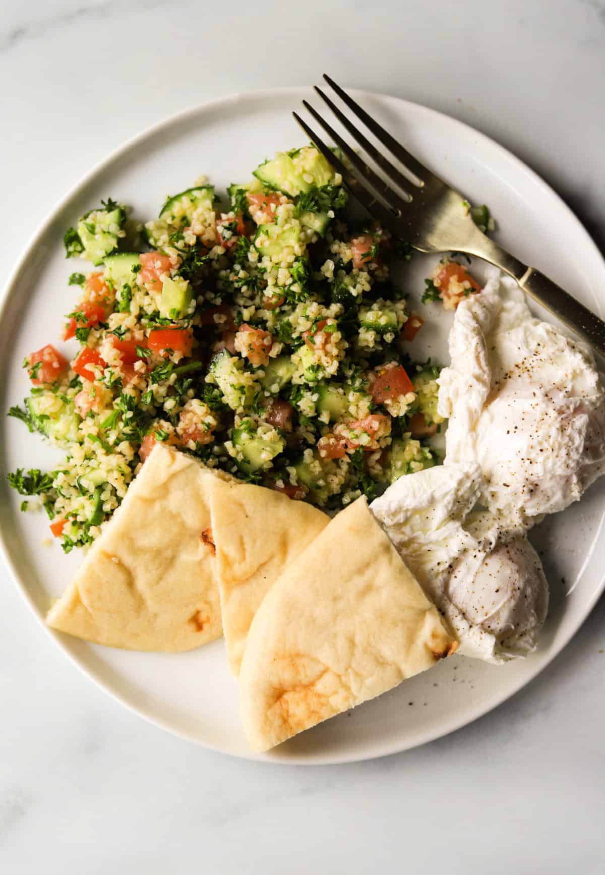 An overhead shot of a plate of tabouli with poached eggs and wedges of pita.