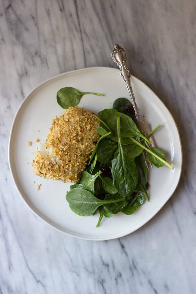 Overhead shot of Pistachio-Crusted Cod on a white plate with fresh spinach leaves. 
