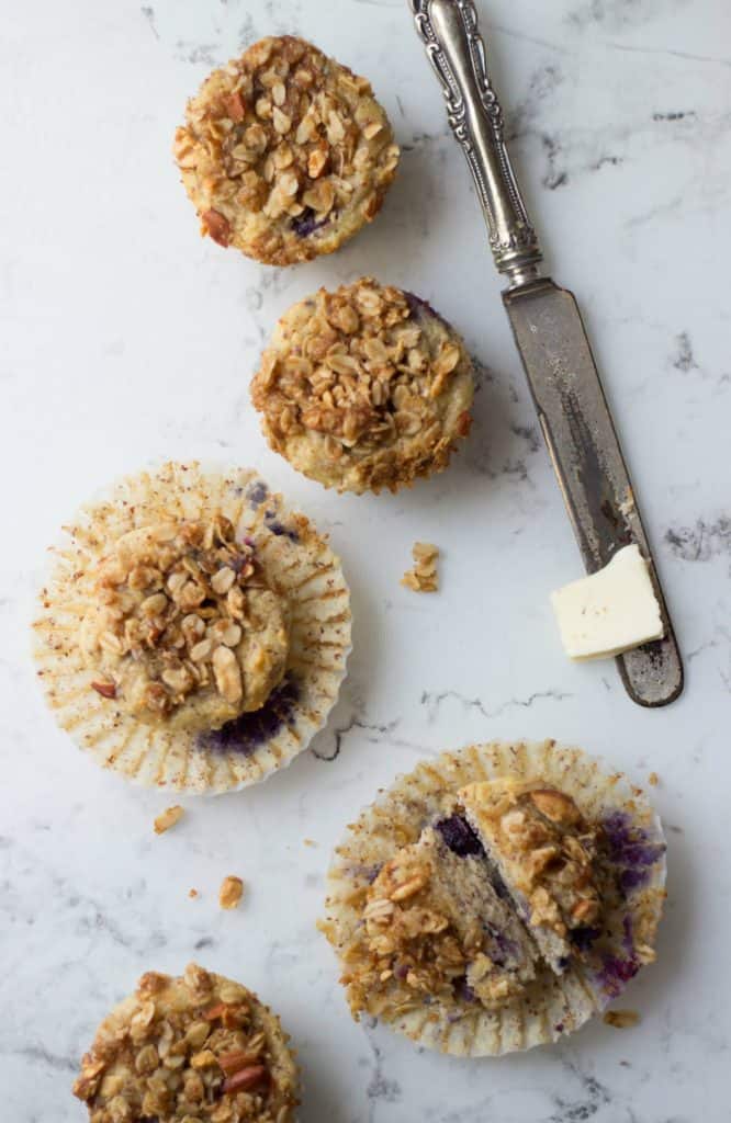 Overhead shot of Almond Crunch Blueberry Muffins on a white counter with a small sliver of button a butter knife. 