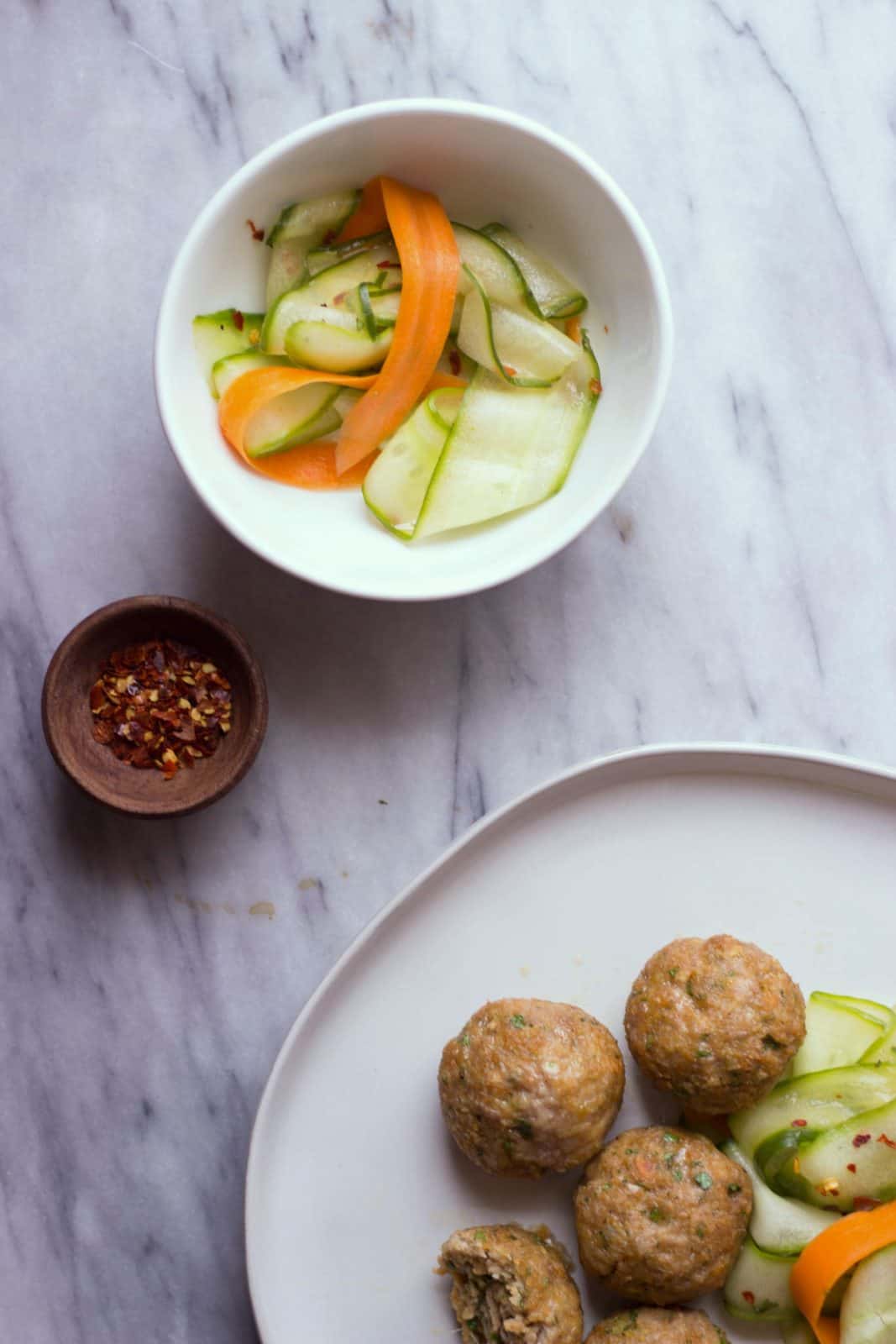 Overhead shot of Asian-Style Pork Meatballs with Sesame Cucumber Salad and a small wooden bowl with red pepper flakes.
