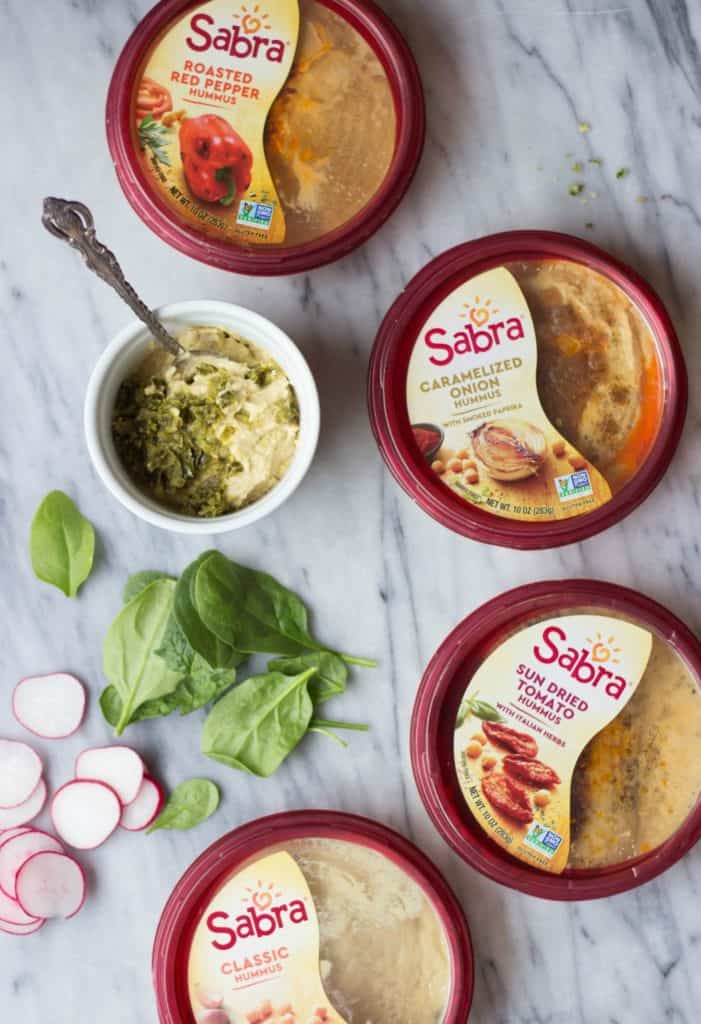 Overhead shot of Sabra hummus containers next to fresh spinach and radishes and hummus in a white bowl. 