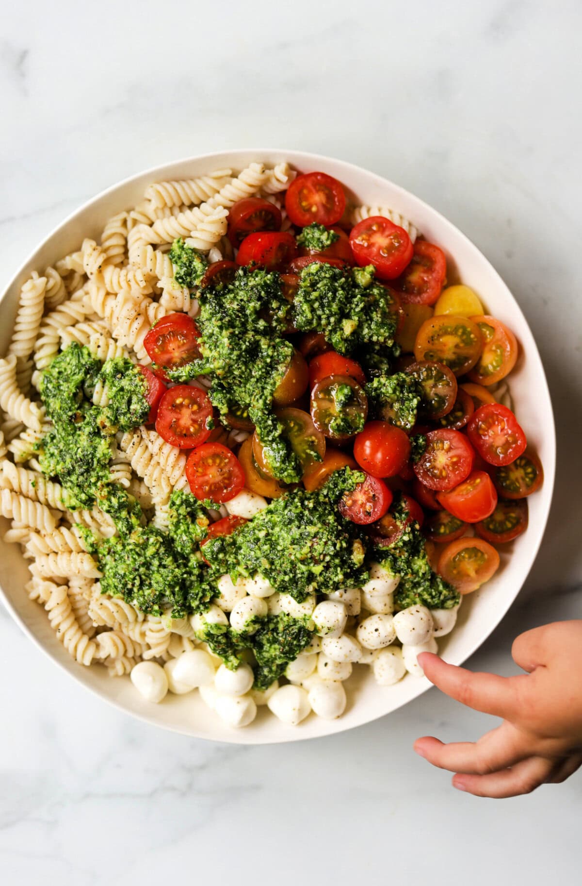 An overhead shot of a bowl of ingredients for pesto pasta salad on a marble backdrop with a hand reaching in.