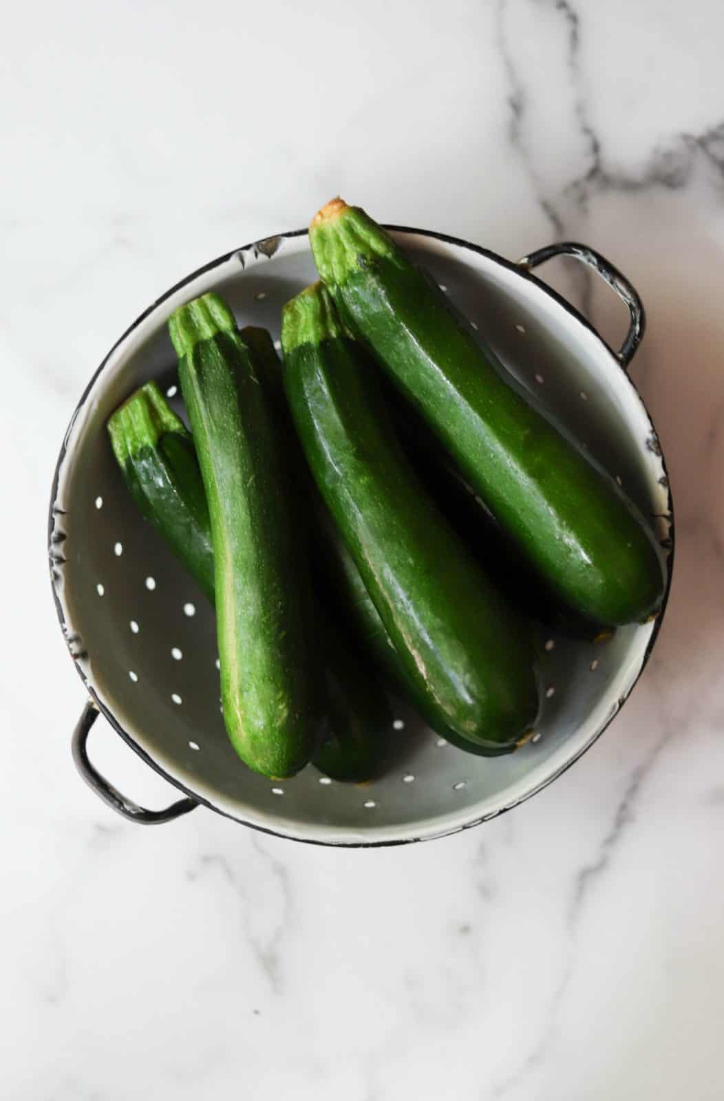 Overhead shot of fresh zucchini in a white colander as an example of an in-season vegetable option to save money on groceries. 