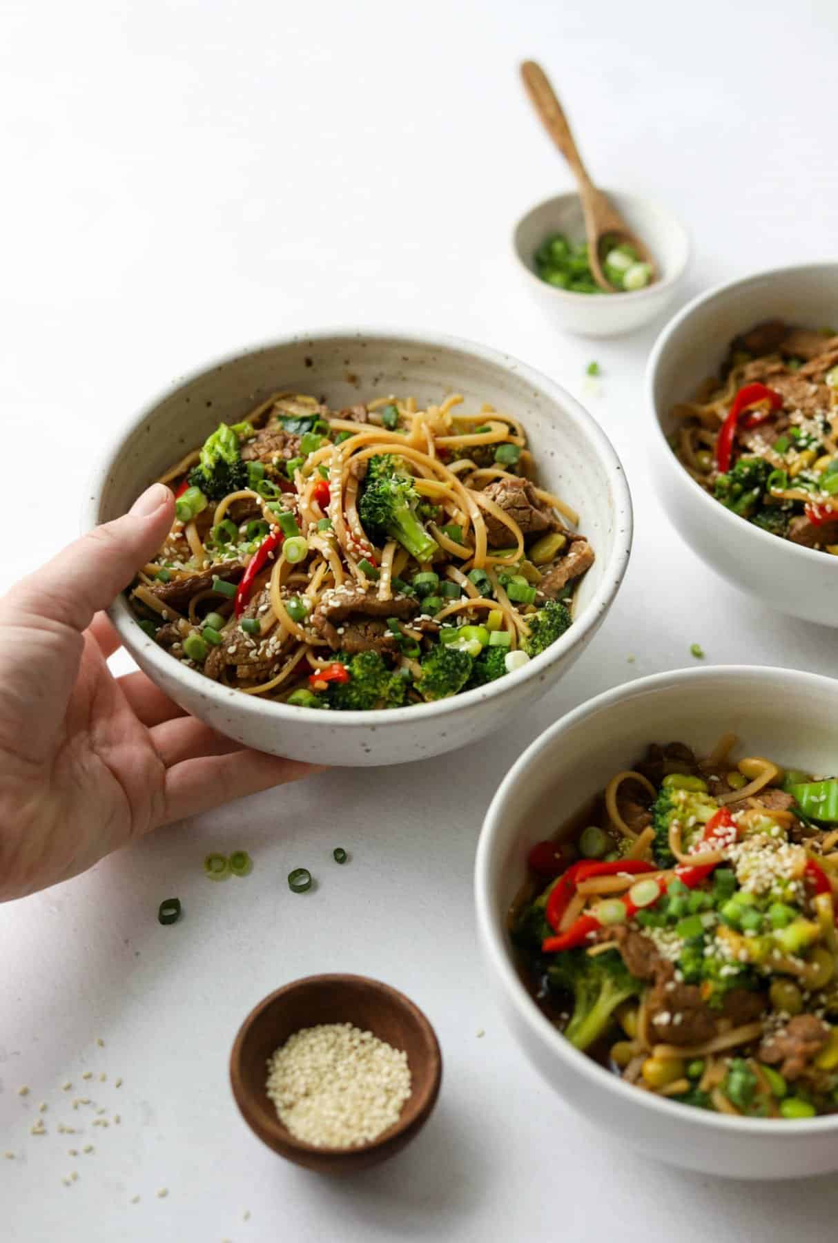 Side shot of Asian-Style Beef & Veggie Noodle Bowls with a hand holding one bowl.