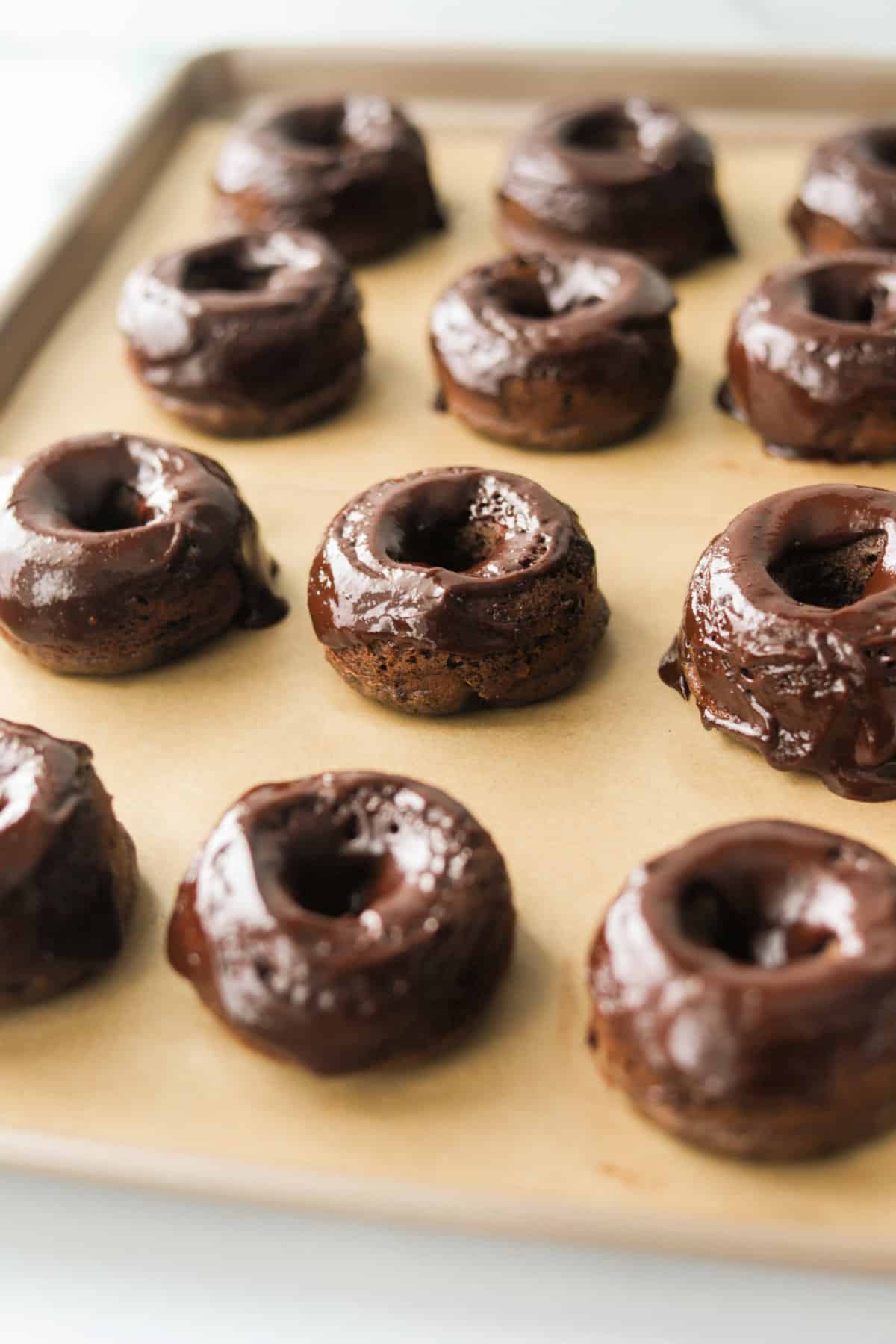 A side angle shot of chocolate donuts lined on a baking sheet.