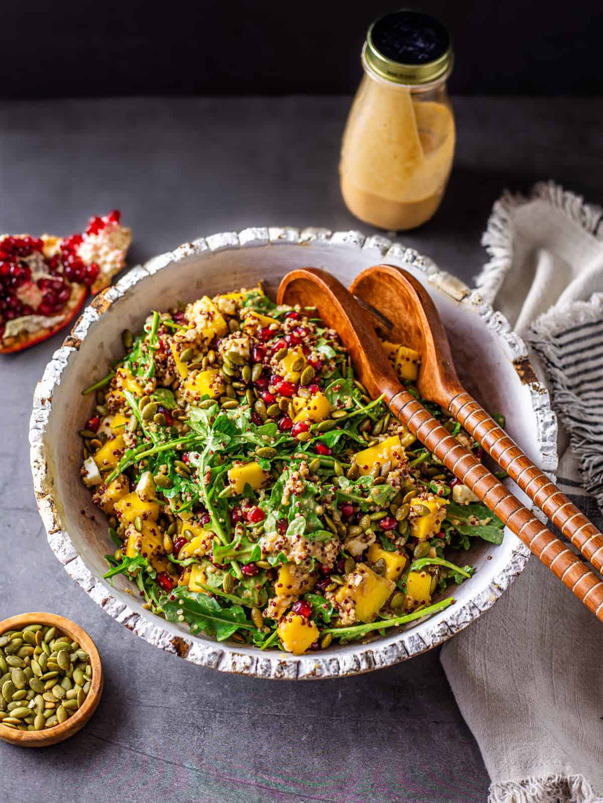 A front shot of a bowl of arugula salad with pepitas, mango, pomegranate arils, queso fresco and pomegranate dressing. Features wooden salad tongs with ingredients in dishes on the side with a kitchen cloth.