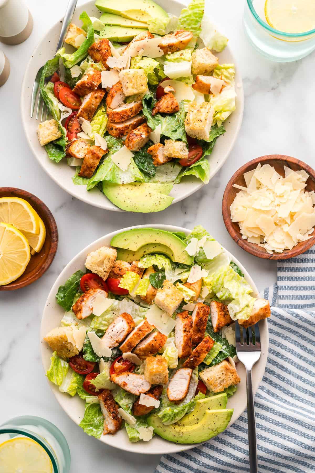 An overhead shot of aplates of chicken caesar salad with lemons and Parmesan in bowls on the side.