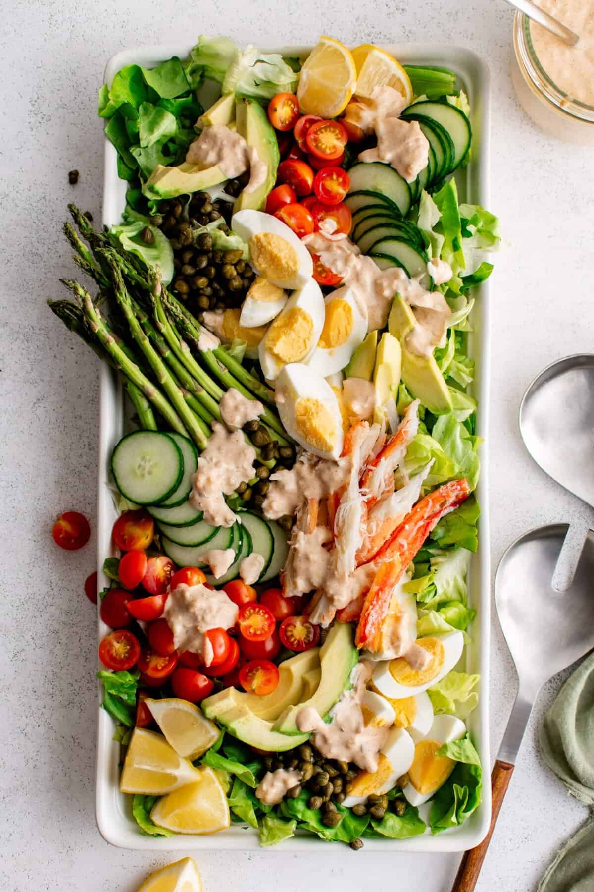 A rectangular platter with crab nicoise salad with asparagus, boiled eggs, capers, cucumber and tomato and a creamy dressing drizzled on top.