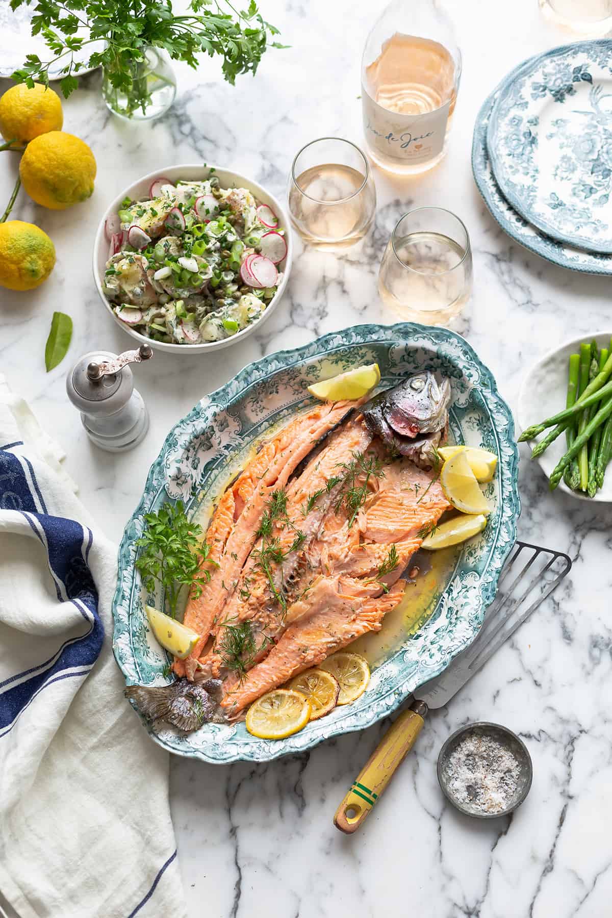 An overhead shot of a whole roasted fish with herbs, lemons, salad and wine surrounding the platter.