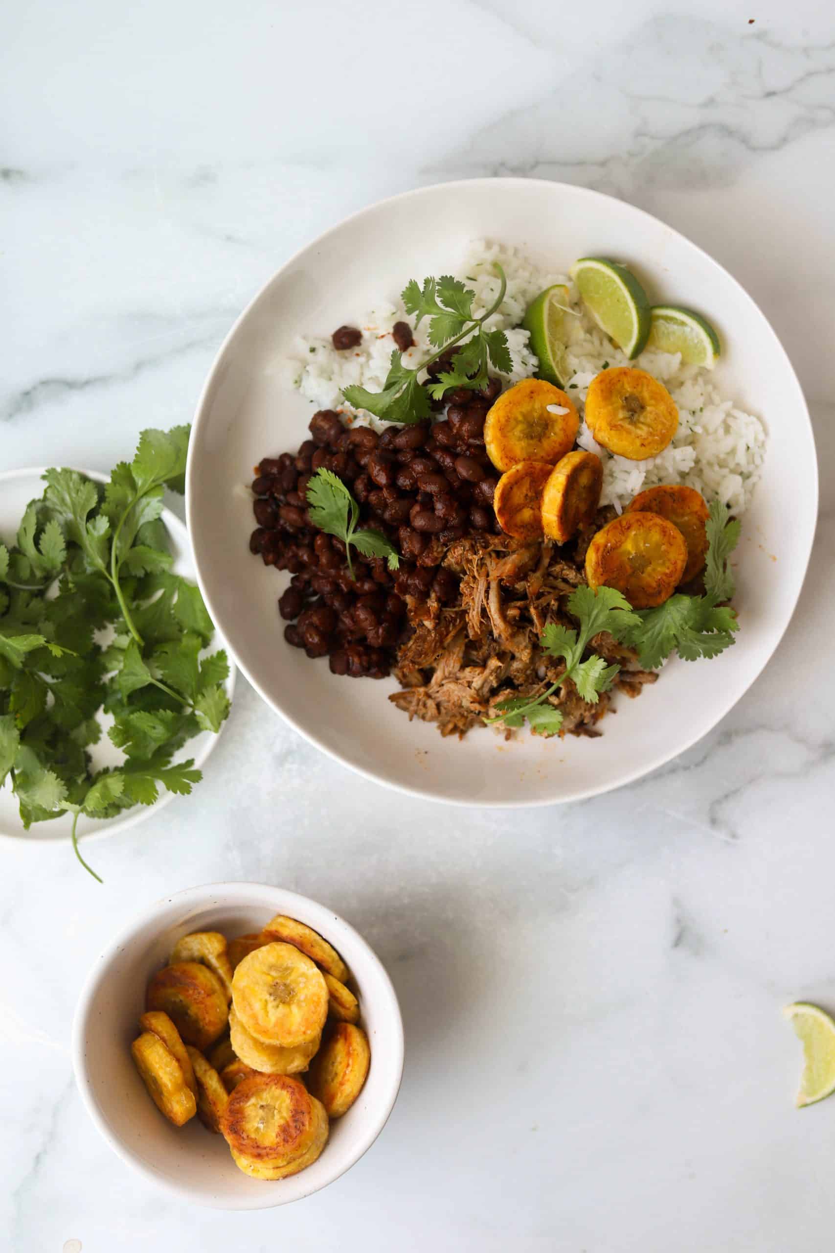 A large white bowl filled with cuban pork, rice, black beans and fried plantains.