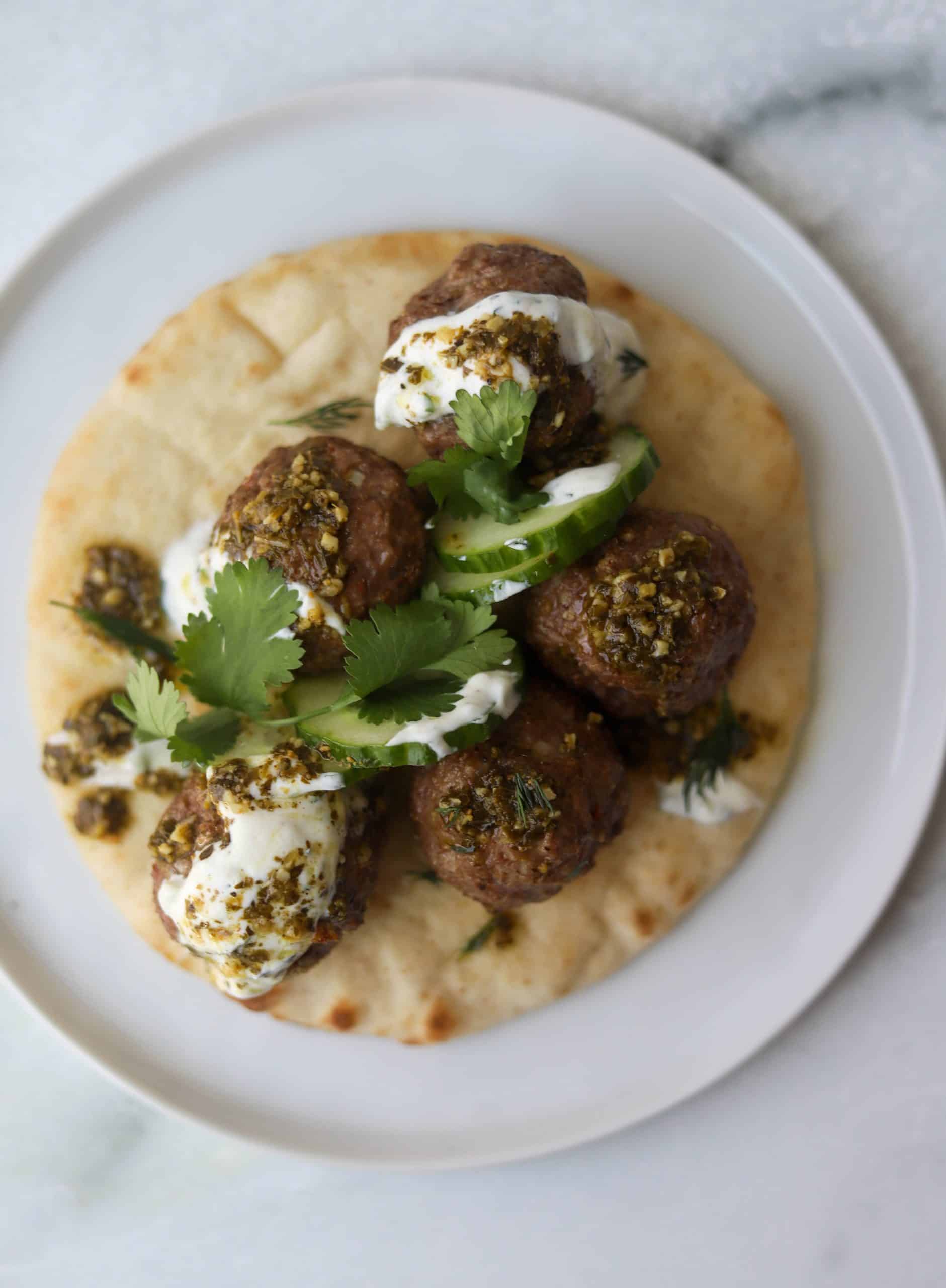 Greek lamb meatballs on top of a pita, topped with cucumbers, dill, parsley, pesto, and tzatziki sauce.