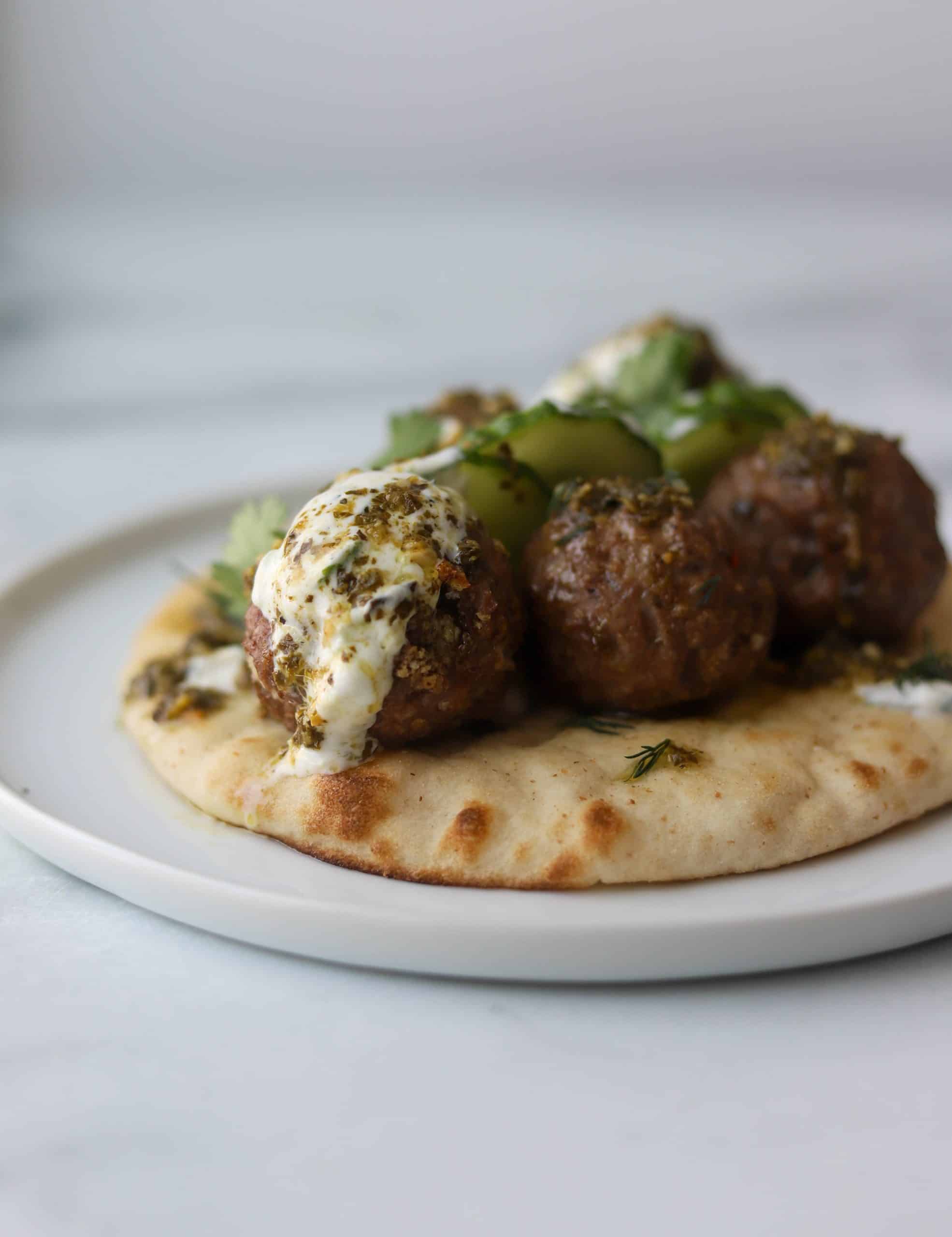 A side view of meatballs topped with tzatziki sauce, pesto, and cucumbers on top of a pita.