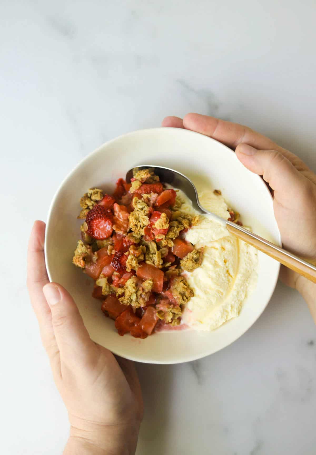 A birds eye view of a hand holding a bowl with strawberry rhubarb crisp with vanilla ice cream.