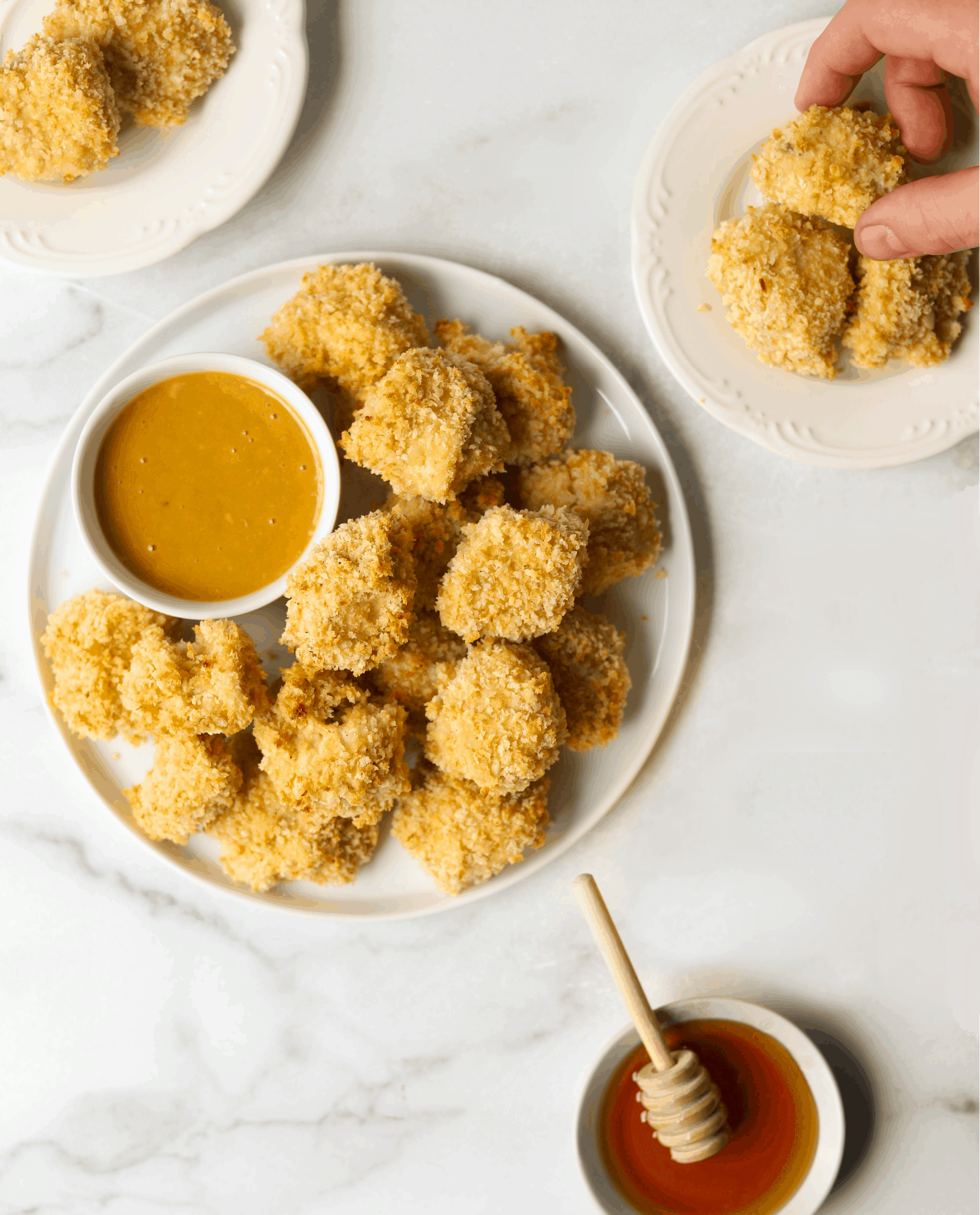 A plate of chicken nuggets on a white marble backdrop with a bowl of honey 
