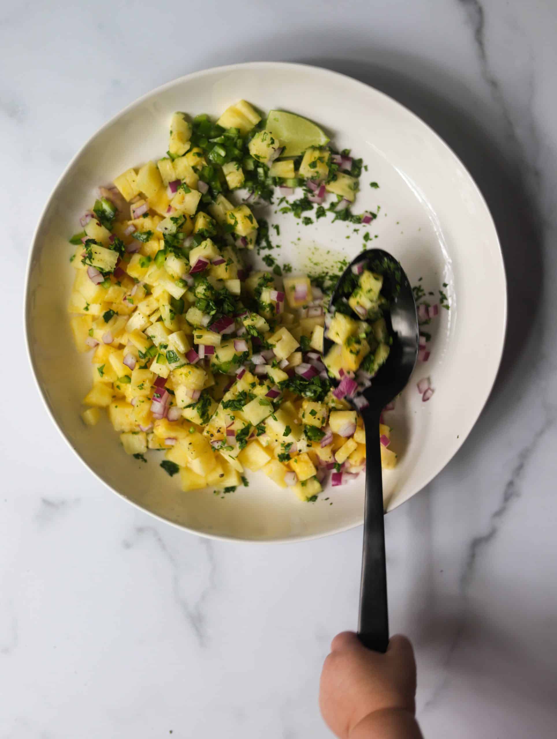 A white bowl with pineapple salsa with a hand holding a spoon.