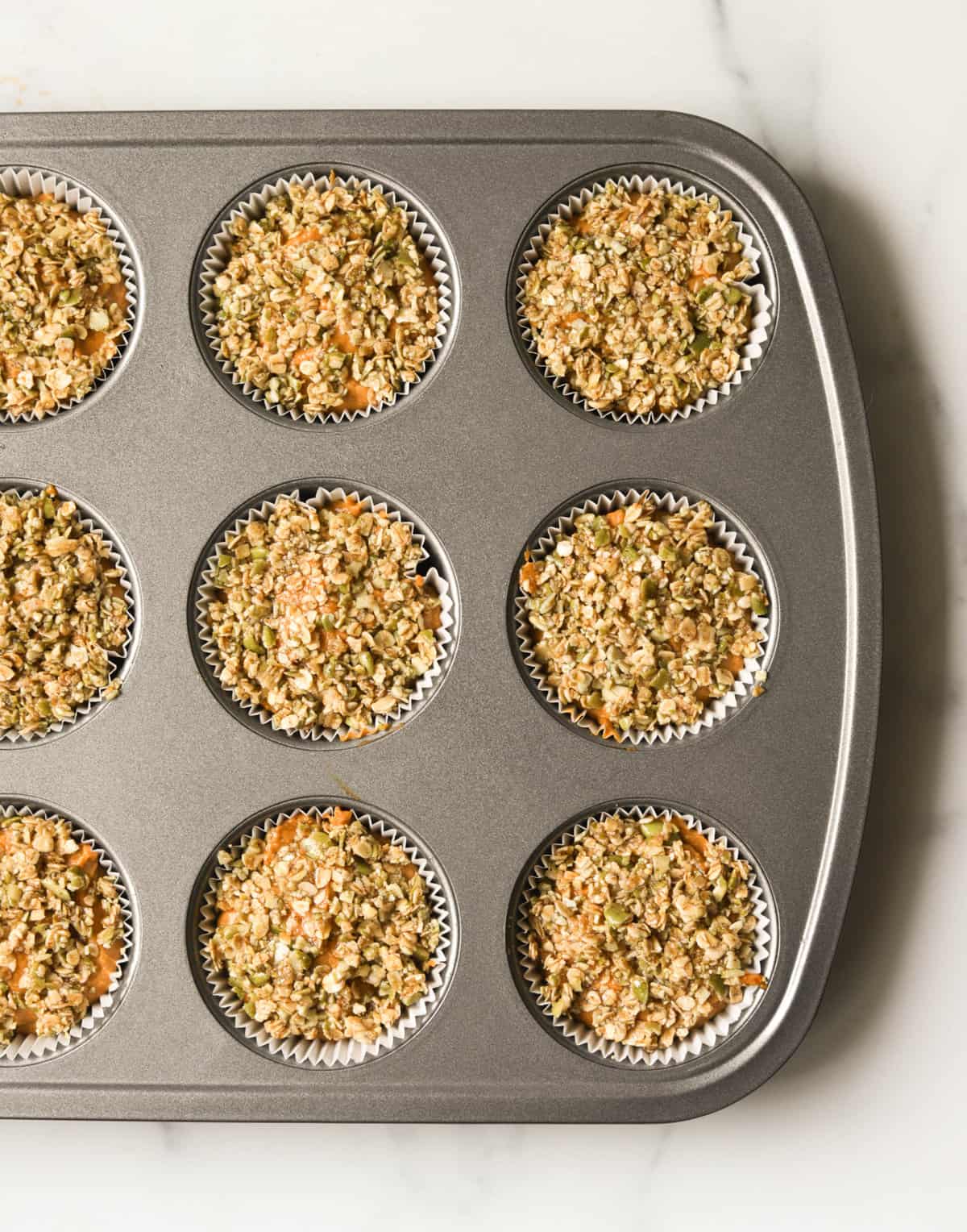 An overhead shot of a muffin tin with oat flour pumpkin muffin batter with pistachio crumble on top.