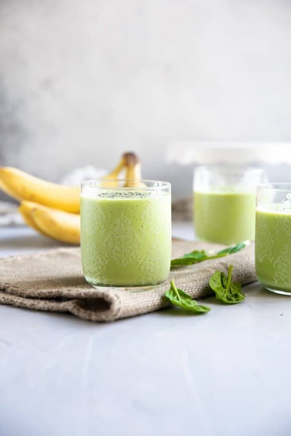 A front view of green smoothies in clear glasses with a bunch of bananas in the background.