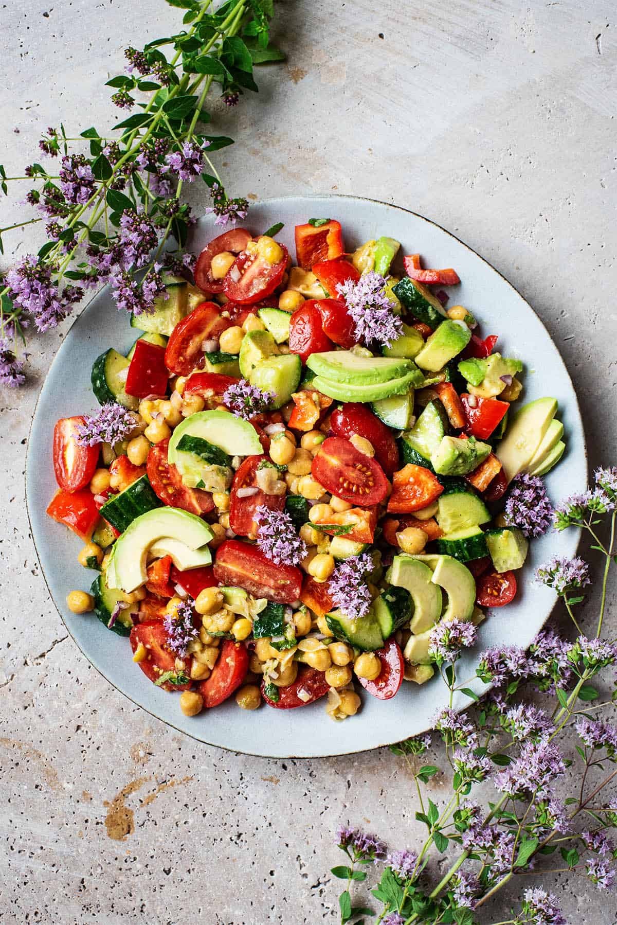 An overhead shot of a plate of chickpea salad with peppers, tomatoes, avocado and cucumber in a Greek dressing. Features purple flowers on the side.