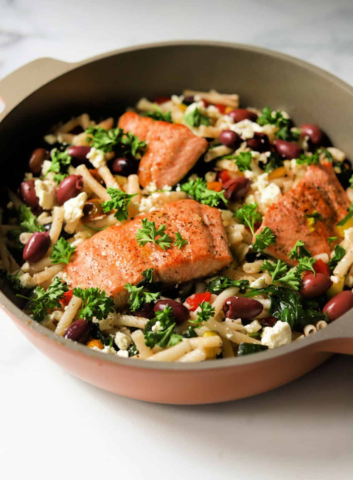 A side shot of a skillet of Mediterranean pasta with salmon, vegetables and herbs.