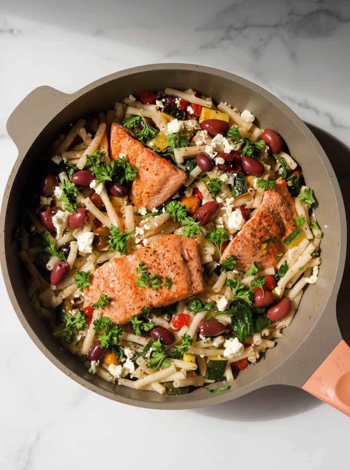 An overhead shot of a pan of lemony pasta with Mediterranean vegetables and herbs and pan-seared salmon on top.