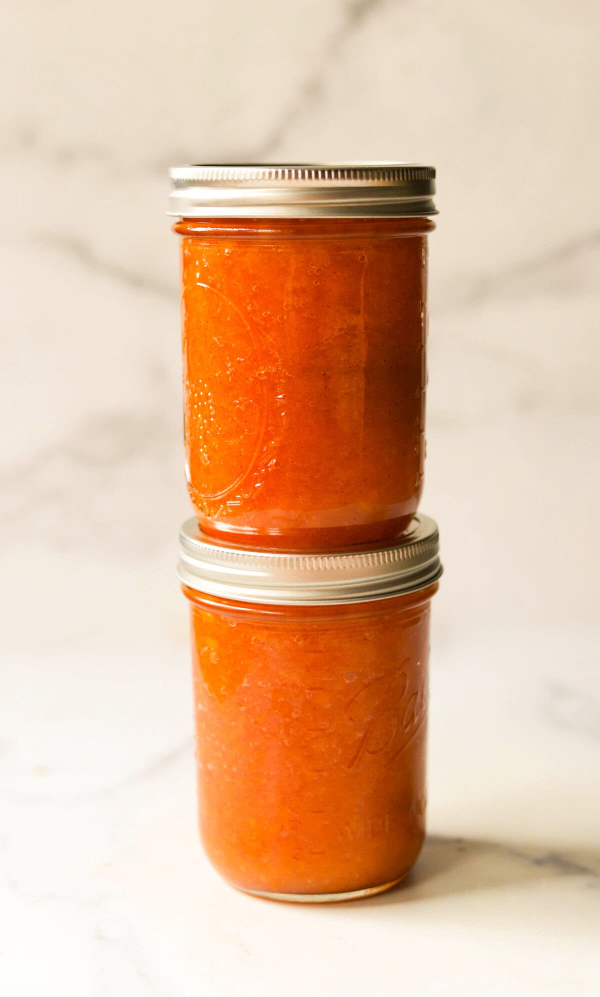 A stack of two jars of fresh peach jam on a marble backdrop.