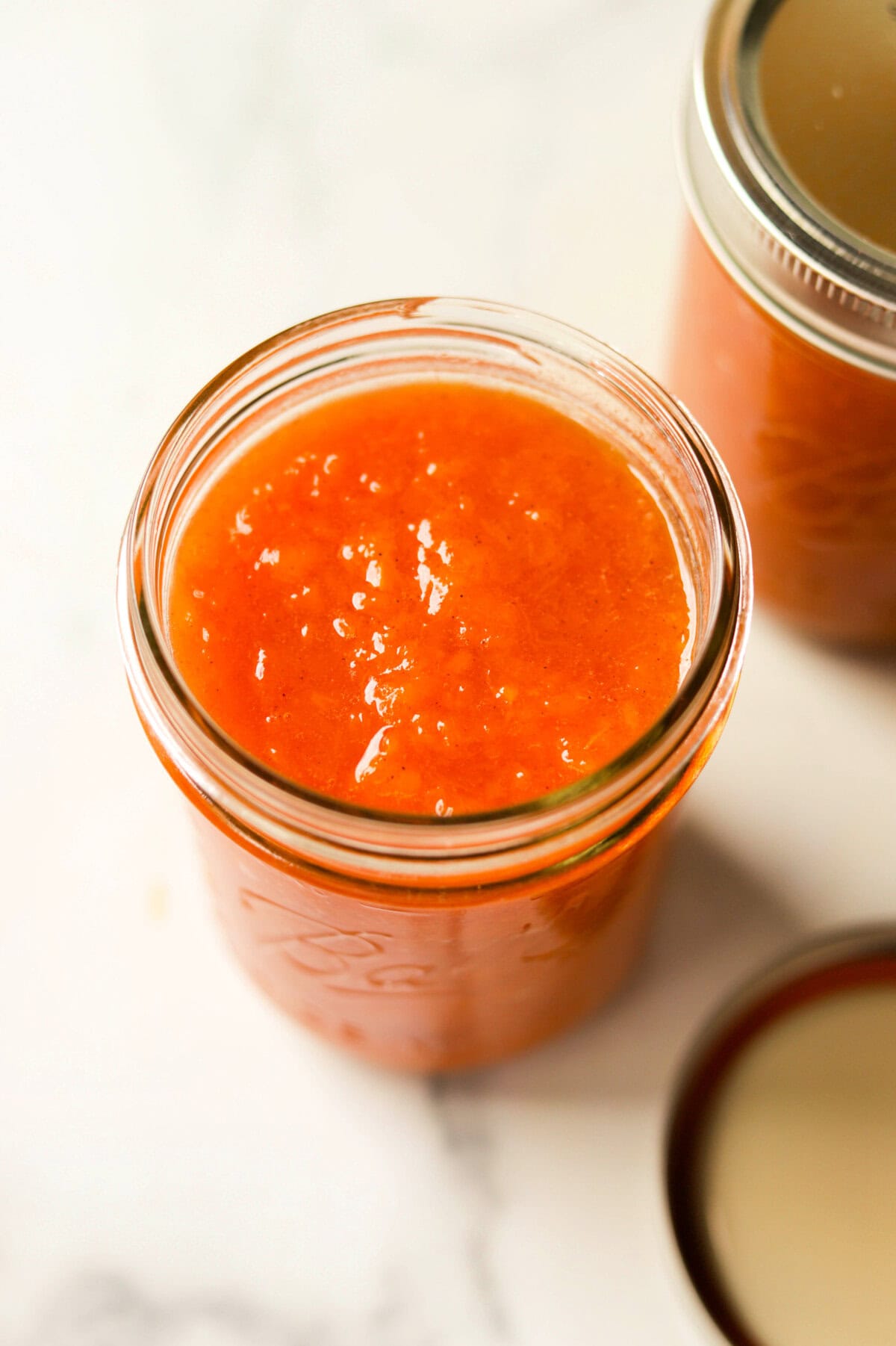 A side angled image of a jar of fresh peach jam with the lid off an a full jar in the backdrop.