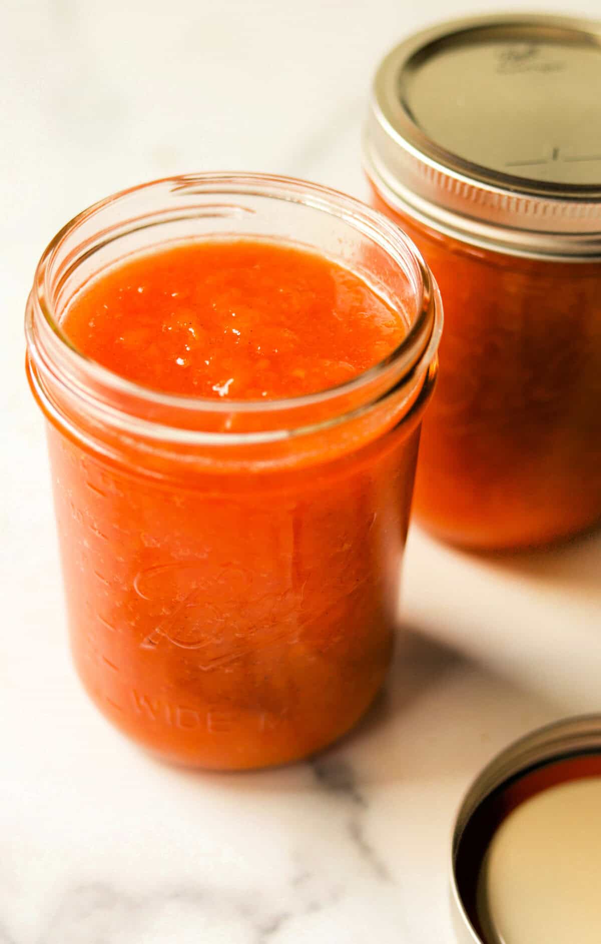 A close up angled image of a jar of fresh peach jam with a full jar in the background and a lid on the marble backdrop.